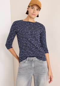 Marineblau und hellblau gestreiftes Langarmoberteil mit rundem Halsausschnitt, kombiniert mit hellgrauen Jeans. Das Oberteil hat einen strukturierten Stoff und einen lässigen Schnitt.