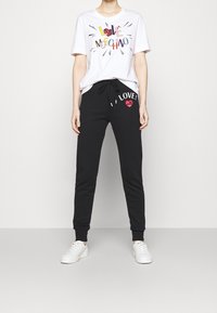 White t-shirt with multicolored graphic "LOVE MOSCHINO" and black sweatpants featuring "I LOVE!" and a red heart print, paired with white shoes.
