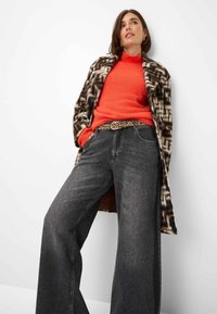 Kunstfellmantel mit einem geometrischen Braun- und Weißmuster, orangem Pullover, Gürtel im Leopardenmuster und locker sitzenden, dunkelgrauen Jeans.