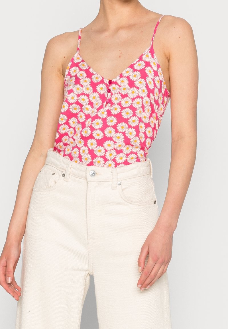 Femme portant un haut à bretelles fines rose avec un imprimé marguerites blanches et jaunes, rentrée dans un pantalon taille haute écru, sur un fond uni.