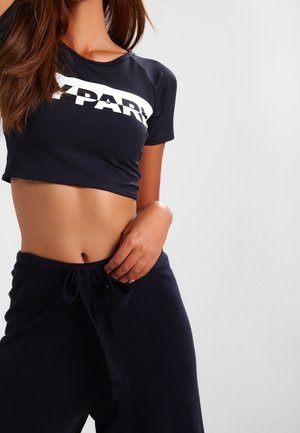Donna che indossa una maglietta sportiva nera accorciata con lettering bianco e pantaloni neri con coulisse, mano posata vicino alla vita, sfondo neutro.