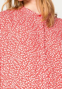 Blouse rouge à motif floral avec de petites fleurs blanches, tissu léger, col rond et détail de boutons à l'avant. Surface texturée au toucher doux.