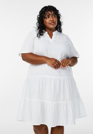 Kobieta o kręconych włosach, ubrana w luźną białą sukienkę z krótkim rękawem i warstwami, patrzy lekko w górę, z rękami delikatnie złożonymi z przodu.