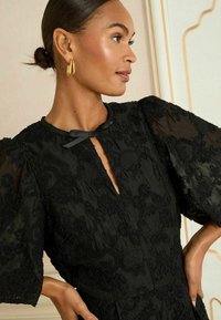 Juoda siuvinėta suknelė su aukštu kaklu, priekyje su išpjova, pūstomis rankovėmis ir satino akcentu. Išsiuvinėtas gėlių raštas visoje suknelėje.