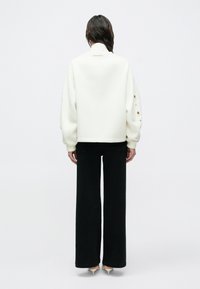 Άτομο που στέκεται με την πλάτη προς τα εμάς φορώντας ένα λευκό μπουφάν με οπές στα μανίκια, μαύρο παντελόνι φαρδύ στην άκρη και λευκά παπούτσια με τακούνι σε ένα απλό φόντο.