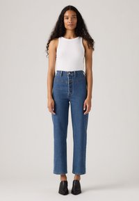 Donna con lunghi capelli ricci che indossa un top bianco senza maniche, jeans blu a vita alta e scarpe nere con punta a stiletto, in piedi di fronte.