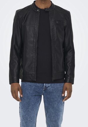 Homme portant une veste en cuir noire avec poche poitrine zippée sur une chemise noire et un jean bleu, sur fond uni.