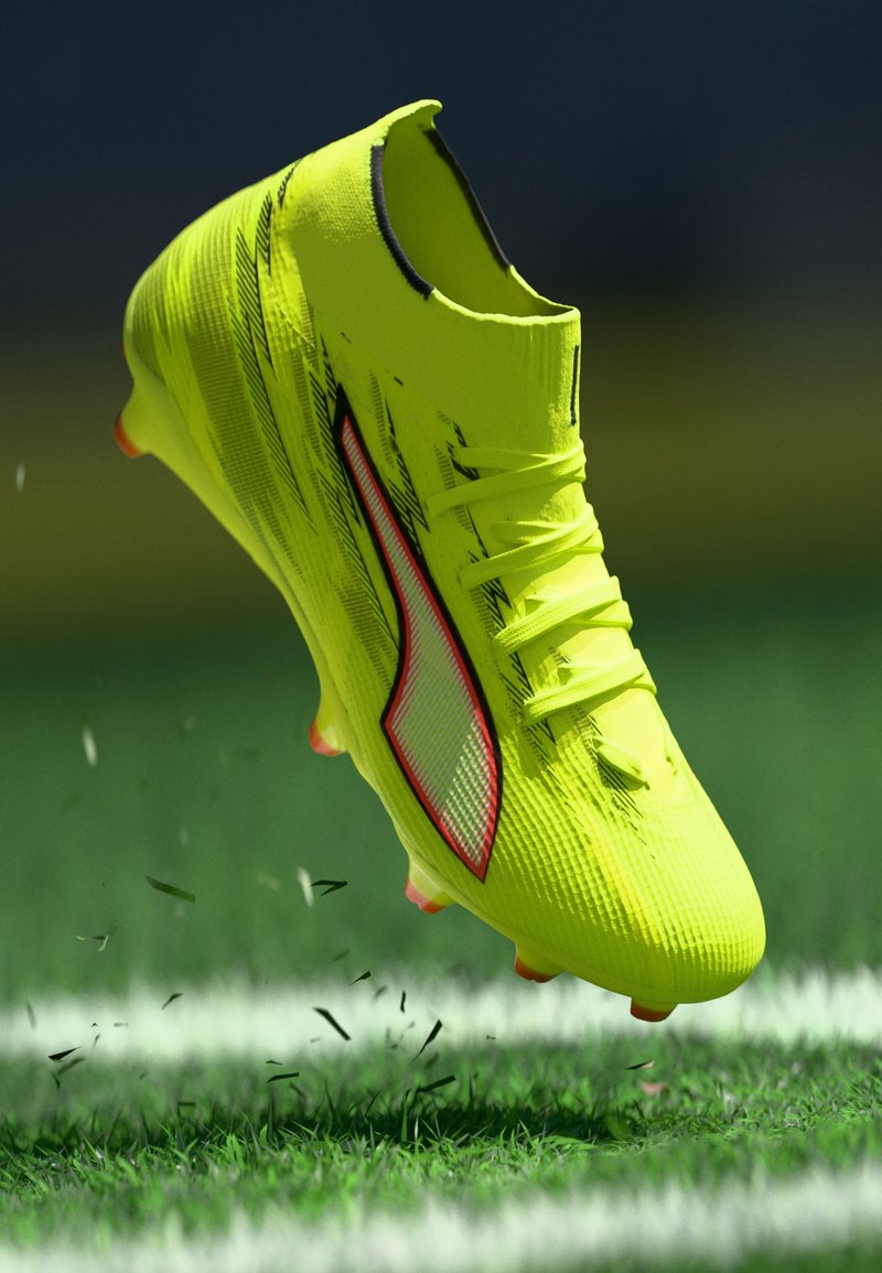 Taco de fútbol amarillo brillante con superficie texturizada, forma aerodinámica, acento rojo y cordones, mostrado sobre césped verde levantando residuos.