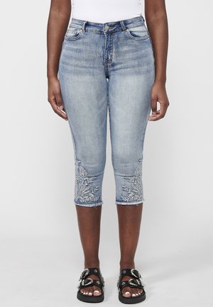 Person iført lyseblå kortere cowboybukser med blomsterbroderi nær kanten, sorte sandaler med spænde og en hvid top.