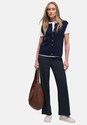 Femme portant un gilet bleu marine boutonné sur un t-shirt blanc, pantalon noir, baskets blanches, tenant un grand sac en cuir marron à ses côtés.