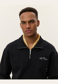 Sudadera negra con cremallera y cuello alto, que presenta texto bordado en blanco "Les Deux" en el pecho y una camisa amarilla con estampado debajo.