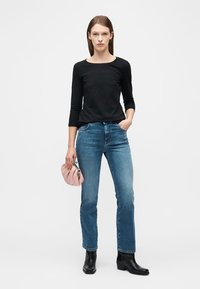 Femme debout portant un haut noire à manches longues, un jean droit bleu, des bottines noires et tenant un petit sac à main rose dans une main.