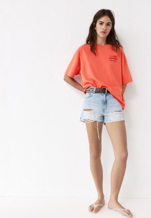 Junge Frau trägt ein oranges „The Sea Lovers“-T-Shirt, zerrissene helle Jeansshorts, einen genieteten Gürtel und weiße Flip-Flops und steht vor einer weißen Wand.