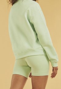 Vaaleanvihreä collegepaita rentolla istuvuudella ja resorireunuksilla, yhdistetty samanvärisiin shortseihin. Pehmeä tekstuuri keltaista taustaa vasten.