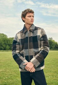 Young man standing outdoors in a blue, gray, and cream checkered zip-up jacket with hands clasped in front, green field and trees behind.