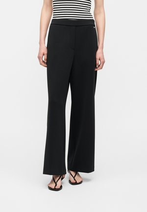 Frau trägt schwarze, weite Hosen, gestreiftes ärmelloses Oberteil und schwarze Zehensandalen, steht vor weißem Hintergrund.