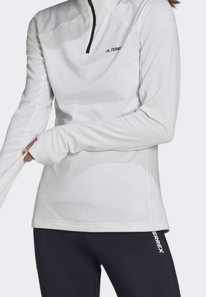 Person justerer tommelfingerhul på hvid langærmet træningstrøje med sort lynlås, kombineret med sorte leggings mærket "TERREX".