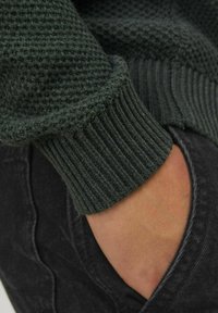 Hand delvis instoppad i en mörkgrå denimficka, iklädd en mörkgrön texturerad stickad tröja med ribbade ärmslut.