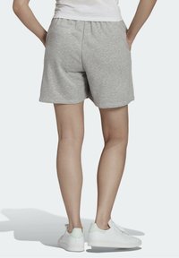 Osoba oblečená v světlých šedých casual šortkách a bílých teniskách, stojící s rukama v kapsách, obrácená zády proti jednobarevnému světlému pozadí.