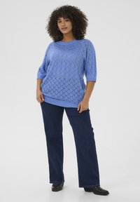 Pull en tricot bleu avec un motif en losanges, à manches courtes et coupe décontractée, associé à un jean évasé bleu foncé et des chaussures noires.