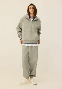 Szary sweatshirt z kapturem i zamkiem, z przednią kieszenią; w zestawie z pasującymi szarymi dresami i białymi trampkami. Posiada paskowany T-shirt pod spodem.