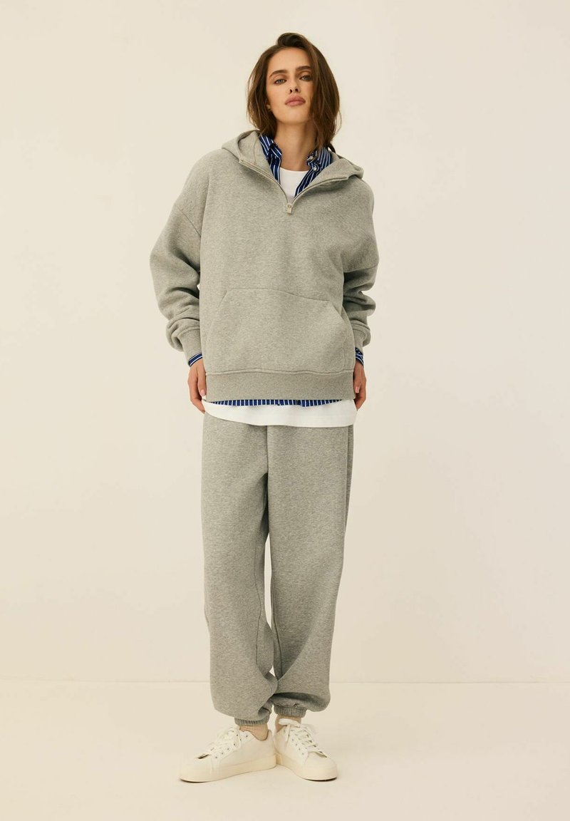 Szary sweatshirt z kapturem i zamkiem, z przednią kieszenią; w zestawie z pasującymi szarymi dresami i białymi trampkami. Posiada paskowany T-shirt pod spodem.