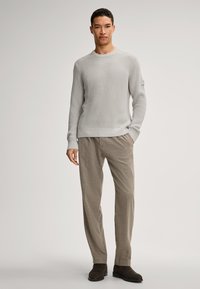 Hellgrauer Rippenstrickpullover mit Rundhalsausschnitt, kombiniert mit beigen, lässigen Hosen und dunkelbraunen Stiefeletten. Einfarbiger Hintergrund.