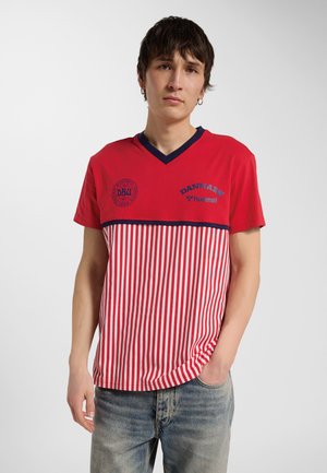 Ung mand med kort mørkt hår iført en rød og hvid stribet dansk sportst-shirt og falmede blå jeans, står med den ene hånd i lommen.