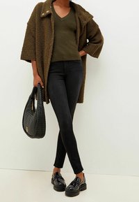 Suéter de punto verde oliva debajo de un abrigo oversized marrón texturizado, combinado con jeans ajustados negros y mocasines negros brillantes. Un bolso de mano negro tejido.