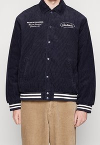 Giacca di velluto a coste blu navy con bottoni a pressione, logo ricamato bianco, tasche laterali e polsini e orlo a righe. Il testo dice "WORK IN PROGRESS."