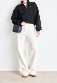 Felpa nera con zip a metà, pantaloni ampia crema, scarpe beige senza lacci e una piccola borsa a tracolla blu navy con logo. Tessuto liscio, vestibilità comoda.