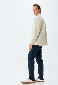Camisa de manga larga beige con bolsillo frontal, combinada con jeans azul oscuro y zapatillas blancas con suelas marrones. Tejido suave, corte relajado.