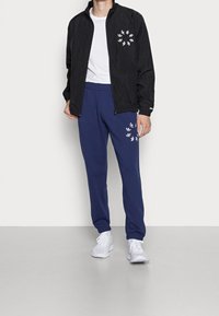 Man i svart zip-up jacka, vit t-shirt, marinblå mjukisbyxor med vita logografiker och vita sneakers som står mot en plain vägg.