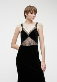 Μαύρο βελούδινο φόρεμα με χαμηλό V ντεκολτέ και δαντελένιες λεπτομέρειες στο μπροστινό μέρος. Μαλακή υφή, εφαρμοστό σχέδιο και ανοιχτή πλάτη.