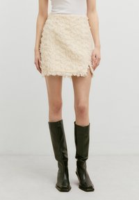 Kurze beige Minirock mit strukturierten Rüschendetails, rohem Saum und seitlichem Schlitz. Kombiniert mit schwarzen knielangen Stiefeln.