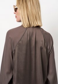 Grijze vloeiende blouse met lange mouwen, zachte textuur en een afgeronde achterbies. Zonnebrillen voegen een chique accent toe.