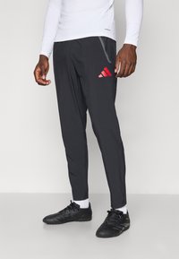 Schwarze Sporthosen mit schmalem Schnitt, mit rotem Adidas-Logo, seitlichen Taschen und Reißverschlussunierschlüssen. Das Material wirkt leicht und strukturiert.