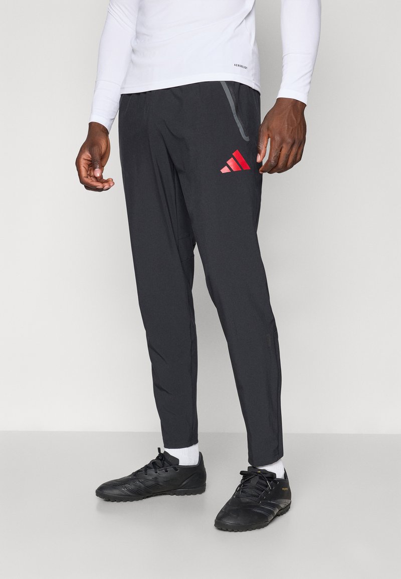 Schwarze Sporthosen mit schmalem Schnitt, mit rotem Adidas-Logo, seitlichen Taschen und Reißverschlussunierschlüssen. Das Material wirkt leicht und strukturiert.