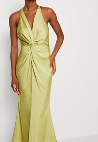 Žena ve světle zelených saténových šatech bez rukávů s detaily zkrouceného uzlu v pase, stojící před jednoduchým bílým pozadím.