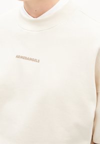 Felpa color crema con un collo alto. Presenta una texture liscia e un logo beige "ARMEDANGELS" sul davanti.