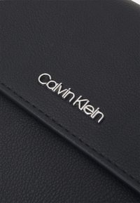Sort tekstureret læderpung med en sølvfarvet "Calvin Klein"-logo. Glat overflade med syninger langs kanterne, ingen synlige mønstre eller detaljer.