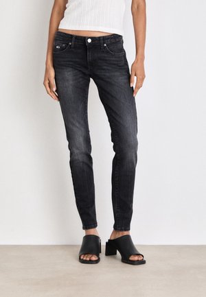 Personne portant un jean noir slim avec un top court blanc et des mules noires à talons ouverts, debout devant un fond uni.