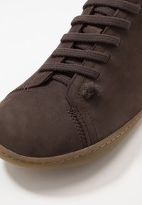 Zapato de ante marrón oscuro con puntera redonda, parte superior texturizada y cordones marrones. Presenta acentos cosidos y una suela de goma ligera y flexible.