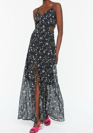 Schwarzes Maxikleid aus transparentem Stoff mit Blumenmuster, tiefem Frontschlitz, Riemchendesign und Cut-out-Details an der Taille. Helle pinke Sandalen.