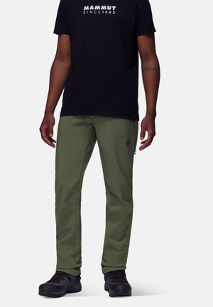 Homme portant un t-shirt noir Mammut, pantalon vert olive avec poche zippée, et chaussures de randonnée noires, debout devant un fond uni.