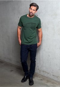 Mörkgrön bomullströja med en grafisk design, kombinerad med mörka jeans och svarta läderstövlar. Bakgrunden är en betongvägg.