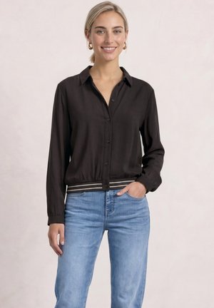 Ung kvinde smiler, iført en sort knappet bluse med stribet kant og lyseblå jeans, stående med den ene hånd i lommen.