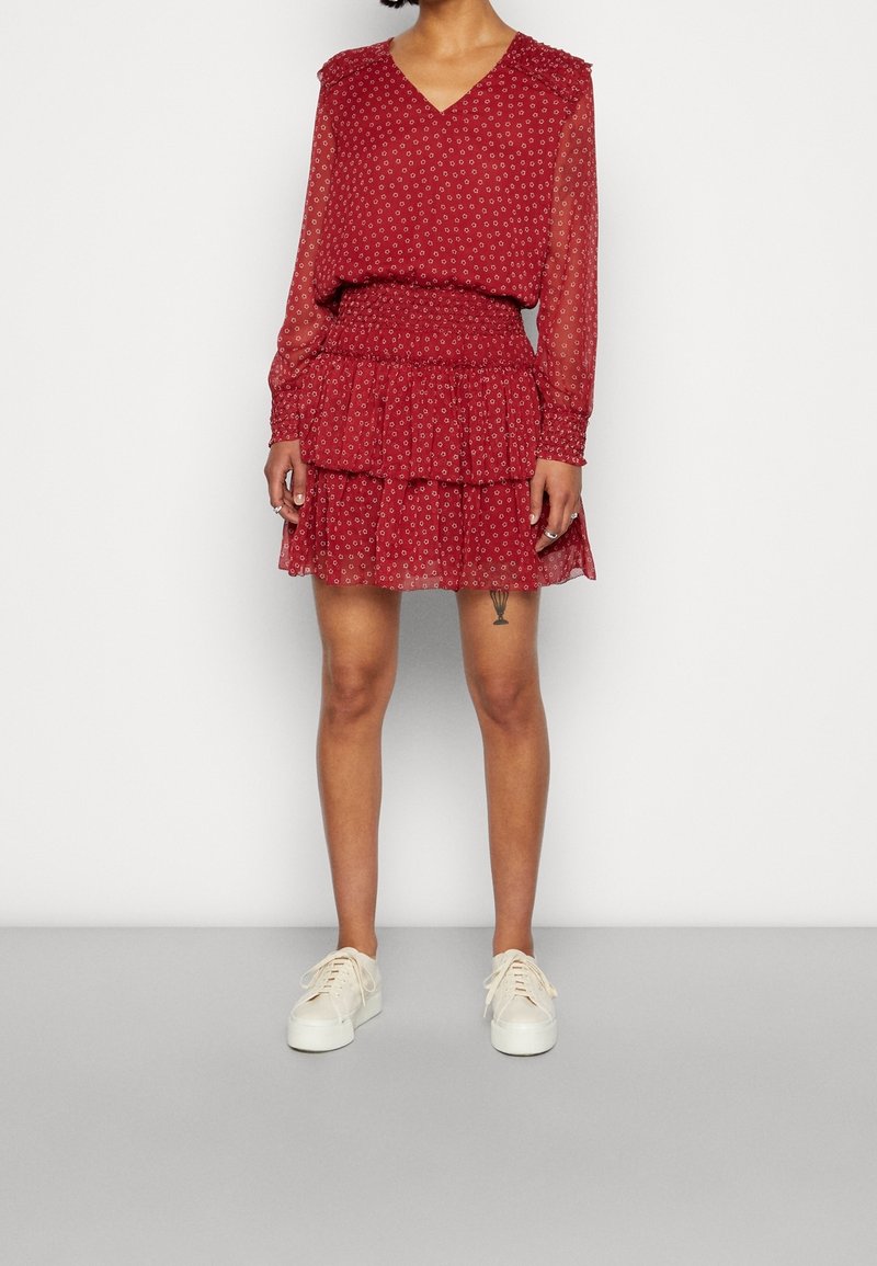 Robe rouge avec un décolleté en V, des manches longues transparentes, une jupe à volants superposés et un petit motif floral. Portée avec des baskets blanches.