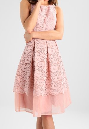 Femme portant une robe rose en dentelle sans manches, longueur genou, avec jupe évasée et doublure transparente, debout les bras croisés et la main près du visage.