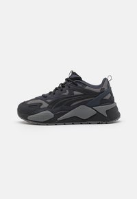 Sneaker atletica nera e grigia con dettagli in rete e suede, suola spessa, chiusura con lacci e linguetta sul tallone, mostrata di lato su sfondo bianco.